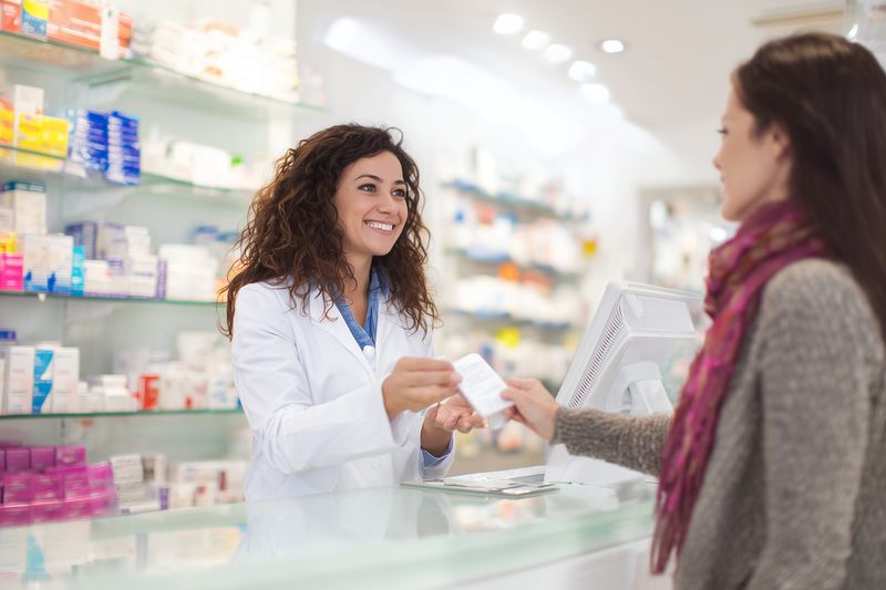 Smiling female pharmacist in white coat hands a medication box to a customer across the pharmacy counter, professional assistance and friendly service in a bright retail healthcare setting.