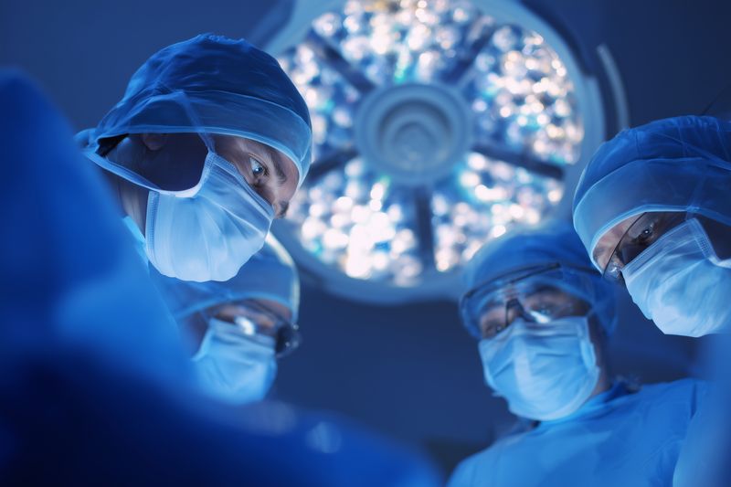 Surgical team in a sterile operating room performing a precise procedure under bright surgical lights, wearing masks, caps and gowns while focusing on patient safety, care and teamwork.