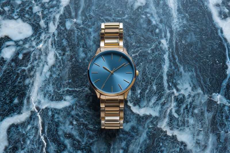 Elegant gold wristwatch with deep blue dial placed on textured blue marble surface, showcasing minimalist timepiece design, metallic bracelet links, polished luxury and refined craftsmanship.