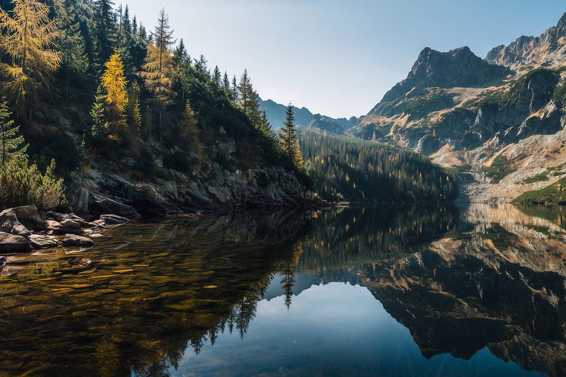 Tranquil alpine lake reflecting rugged mountain peaks and dense conifer forest beneath a clear blue sky, with rocky shoreline and golden autumn foliage creating a serene natural scene.