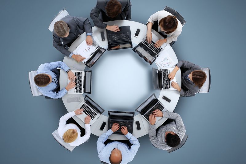 Top view of professionals seated around a circular table working on laptops during a collaborative meeting in a modern office environment, focused on teamwork and productivity.