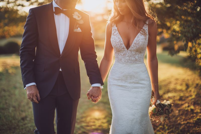 A bride and groom stroll hand in hand through a sunlit field during golden hour, wearing elegant wedding attire and sharing a tender romantic moment with warm backlit glow.