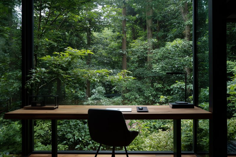 Minimal modern workspace with desk and chair facing a large window that reveals lush green forest, creating a peaceful minimalist home office interior and nature view