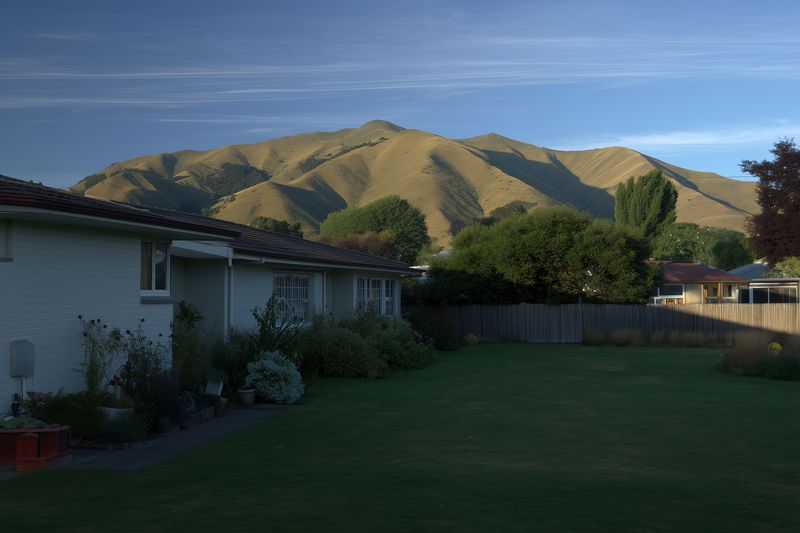 Suburban backyard with manicured lawn, single story houses and garden beds in foreground, rolling sunlit hills and clear sky forming a peaceful residential landscape scene.