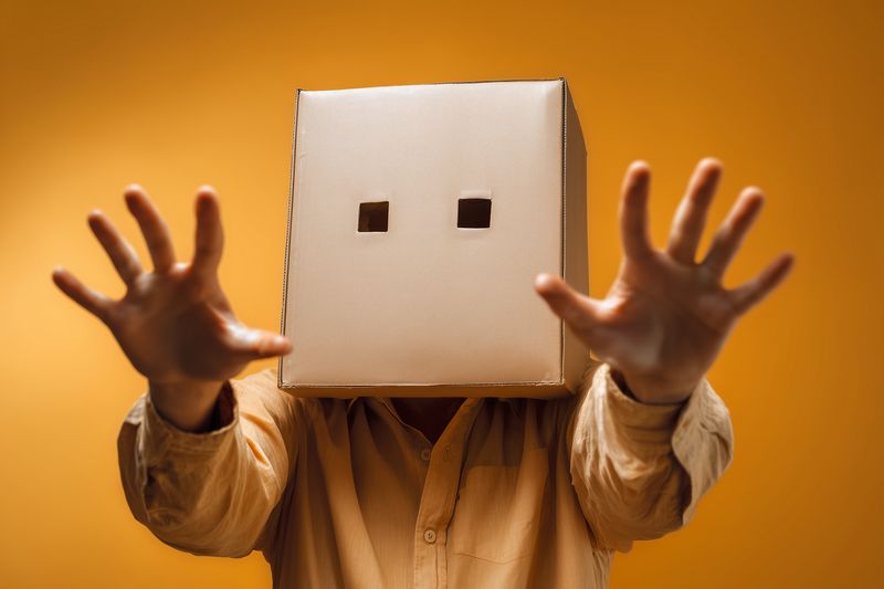 Person wearing a cardboard box with eye holes reaches forward against a warm orange background, conveying mystery, anonymity, playful disguise and creative expression.