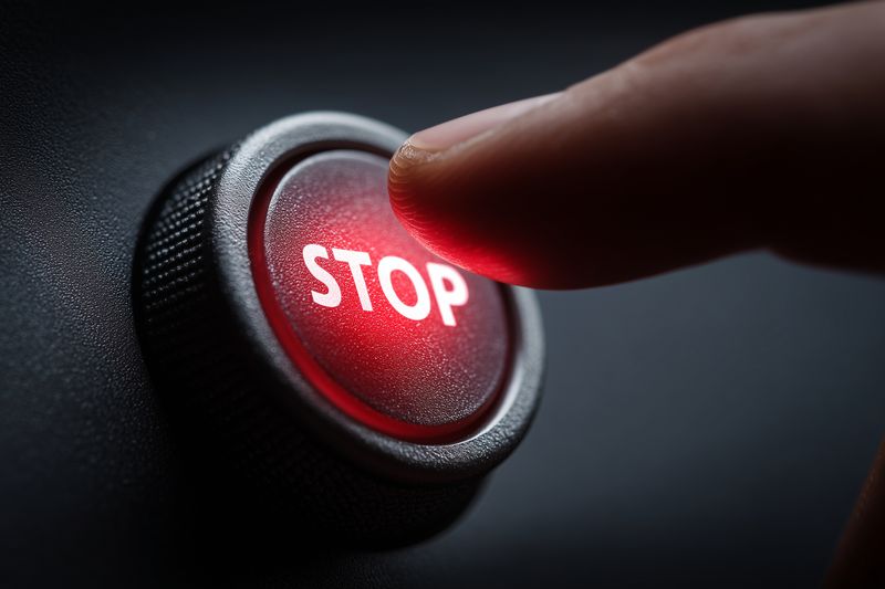 Close-up of a fingertip pressing a red STOP button on a dark control panel, dramatic lighting highlights texture and urgency, evoking emergency shutdown and safety action.