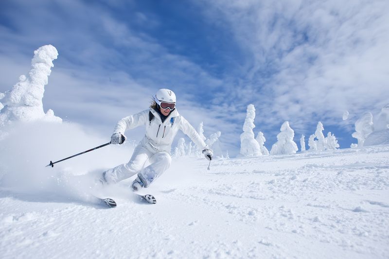 Dynamic action shot of a skier in white gear carving fresh powder on a snowy slope beneath a bright blue sky, surrounded by sculpted snow-covered trees and crisp winter light.