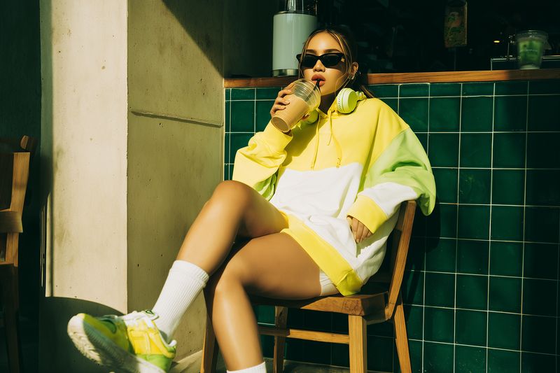 A young woman in an oversized yellow and green hoodie and sneakers sits at an outdoor cafe, sipping an iced drink through a straw while wearing sunglasses and headphones.