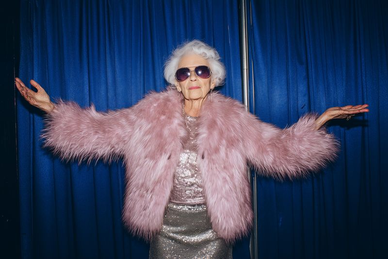 Elderly woman in a fluffy pink faux fur coat and glittering silver dress poses confidently with outstretched arms and sunglasses against a deep blue curtain backdrop.
