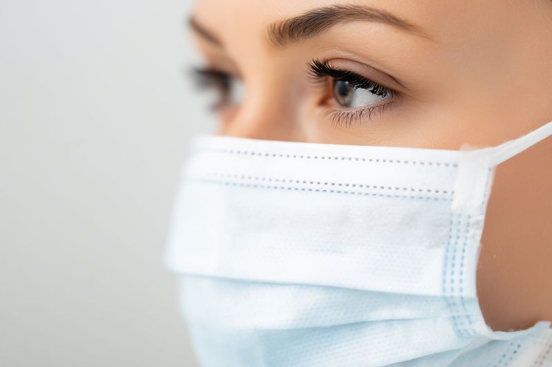 Close-up portrait of a young woman wearing a disposable surgical face mask, focusing on expressive brown eyes and natural makeup, symbolizing health awareness and cautious protection.