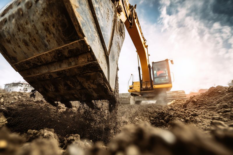 Close-up of an excavator bucket scooping earth on a construction site, backlit by dramatic sky, capturing motion and dirt spray while showcasing heavy machinery power and industrial excavation