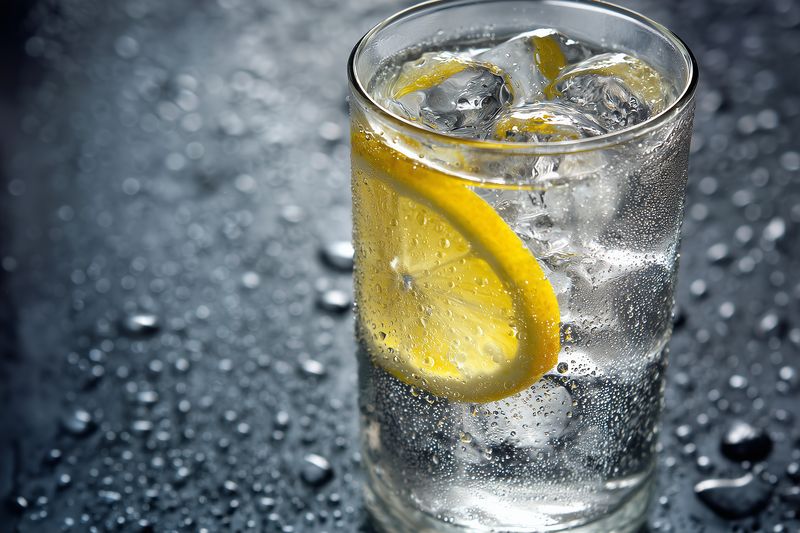 Close-up of a chilled glass of sparkling water with ice cubes and a lemon slice, condensation on the glass and droplets on a dark surface conveying a refreshing beverage moment.