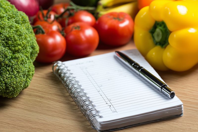 Spiral notebook with pen placed on a wooden countertop surrounded by fresh vegetables including tomatoes, pepper and broccoli, suggesting meal planning and grocery notes.