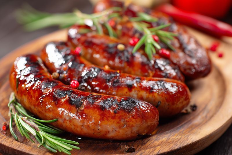 Closeup of grilled sausages on a wooden board, glazed and charred, garnished with fresh rosemary and red peppercorns, evoking savory barbecue and rustic comfort food.
