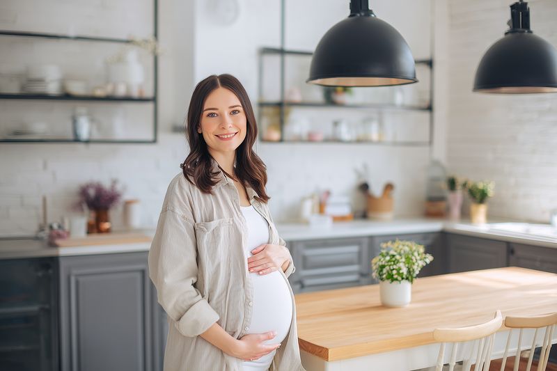 Pregnant woman standing in a bright modern kitchen smiling and gently cradling her belly, conveying warmth, anticipation and maternal joy in a cozy home environment.