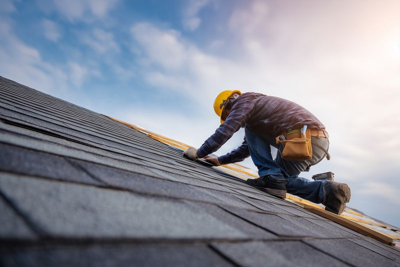 A professional roofer kneels on a sloped shingle roof measuring and aligning materials while wearing safety gear, toolbelt and hard hat under a bright sky.