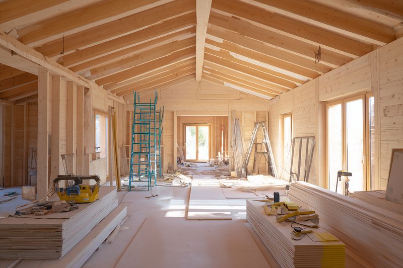 Interior of a wooden house under construction with exposed framing, stacked drywall, ladders and construction tools, bathed in natural light through large window openings.