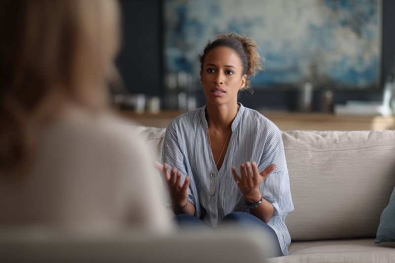 Young woman engaged in an emotional conversation with a counselor on a sofa, expressing concern and openness in a calm living room setting focused on communication and support.