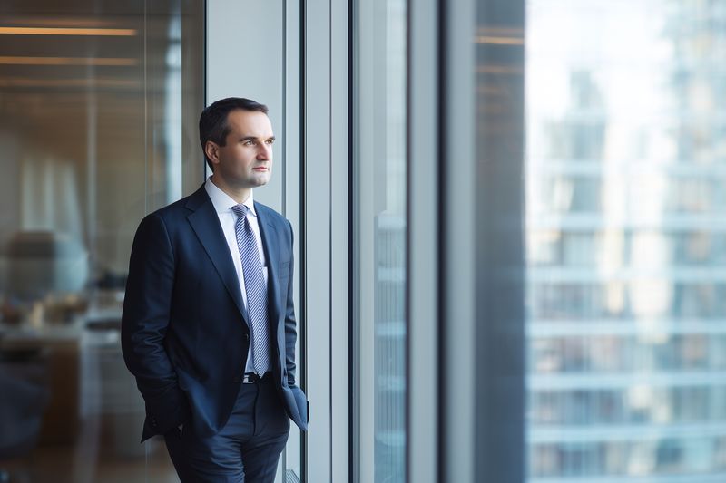 Confident businessman in a suit stands by a large office window looking out thoughtfully, reflecting on work and opportunity in a modern corporate high-rise environment.