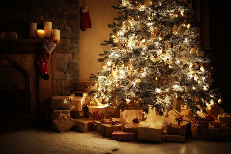 Warm glowing Christmas tree with sparkling lights and ornaments surrounded by wrapped gifts and a cozy fireplace setting, creating a festive intimate holiday atmosphere indoors.