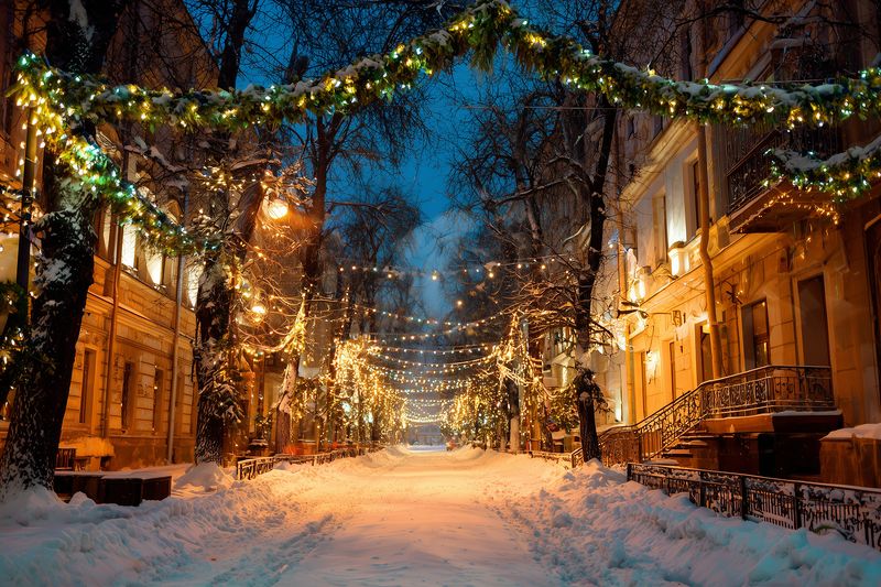 Snowy city street at dusk illuminated by warm string lights and garlands hanging between trees and historic buildings, creating a cozy festive winter evening atmosphere.