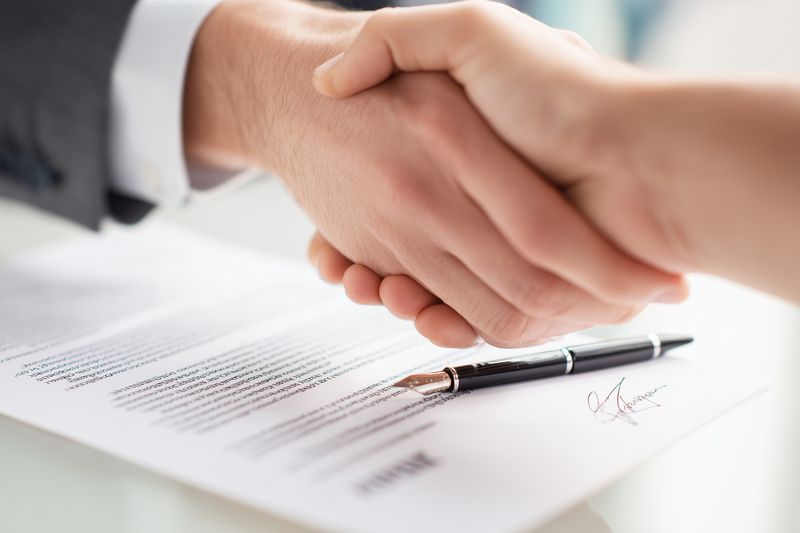 Two professionals shaking hands over a signed contract on a desk, with a pen nearby, symbolizing agreement, partnership, business deal and negotiation in a corporate setting.