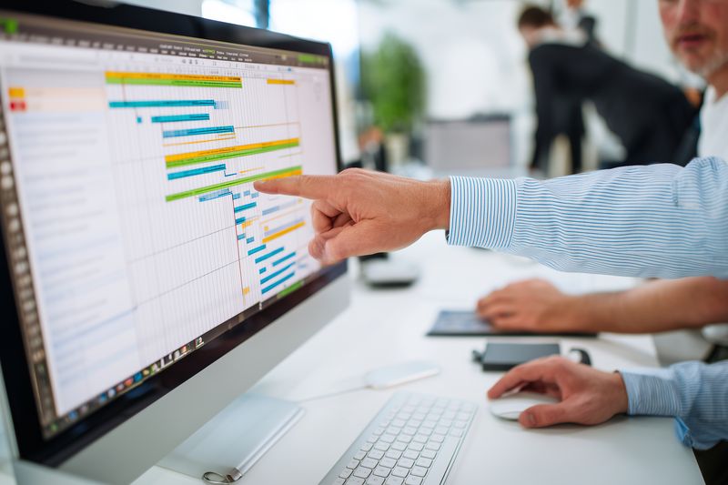 Office colleagues analyze a colorful project timeline on a desktop monitor, pointing at bars and data while planning tasks, schedules and deadlines during a collaborative meeting.