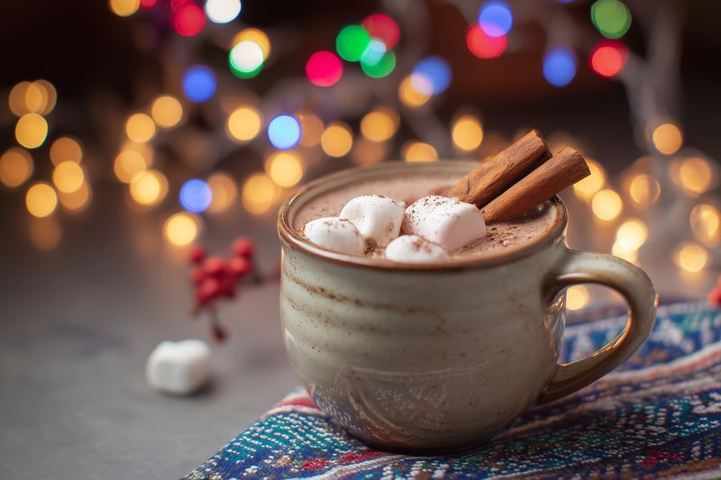 Closeup of a creamy hot chocolate in a ceramic mug topped with marshmallows and cinnamon sticks, set on a patterned cloth with warm festive bokeh lights in the background.