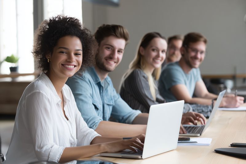 Smiling diverse team of young professionals working together at a bright coworking table, using laptops and notebooks, collaborating in a modern open office during a training session or meeting.