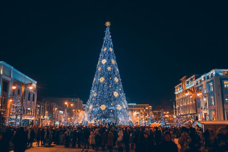 A towering illuminated Christmas tree stands in a bustling city square at night, surrounded by festive lights and a crowd of people enjoying holiday market atmosphere with seasonal decorations.