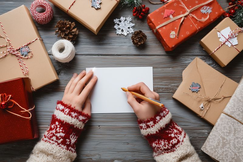 Top view of hands in a cozy fair isle sweater writing a holiday greeting card on blank paper surrounded by wrapped gifts, twine, pinecones and festive decorations on a rustic wooden table.