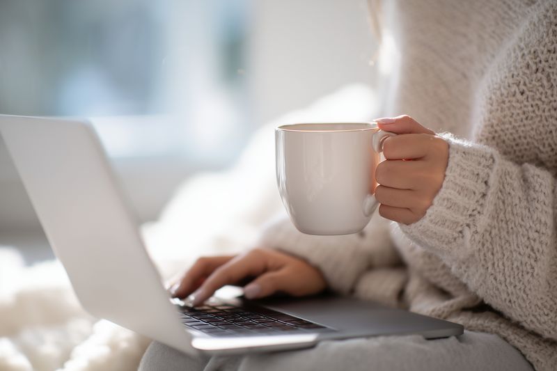 Cozy scene of a person in a soft sweater holding a warm mug while using a laptop, capturing relaxed remote work atmosphere and comfortable home lifestyle moment.
