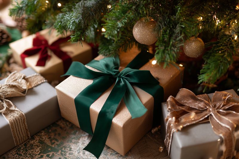Wrapped holiday gifts with elegant ribbons and bows arranged under a decorated evergreen tree with glowing lights and gold ornaments, creating a warm and cozy festive scene.