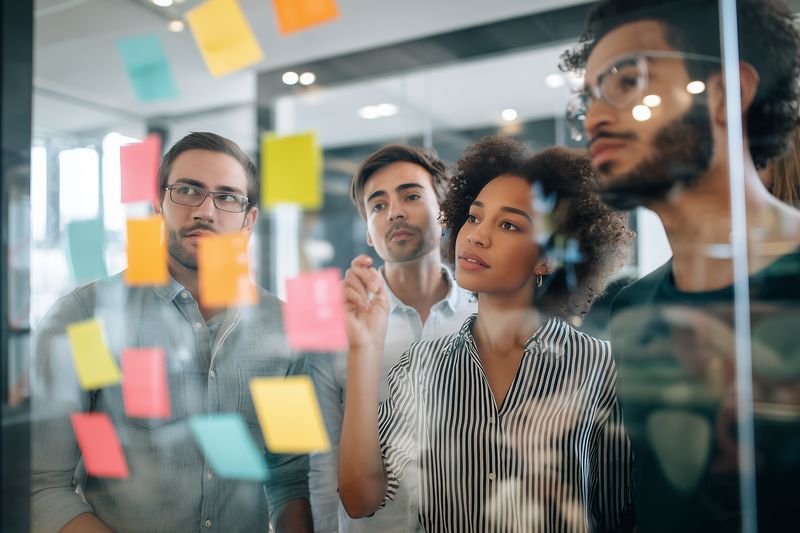Diverse team of professionals collaborating around a glass wall covered with colorful sticky notes, brainstorming ideas and planning strategy during an office creative workshop session.
