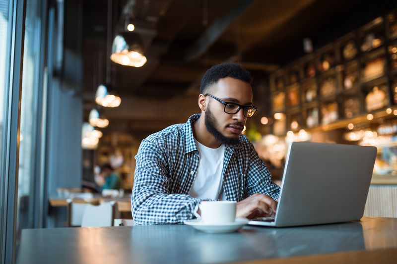 Young man with glasses and beard working on a laptop in a cozy cafe, focused and typing while drinking coffee. Warm ambient lighting and modern interior suggest productive work.
