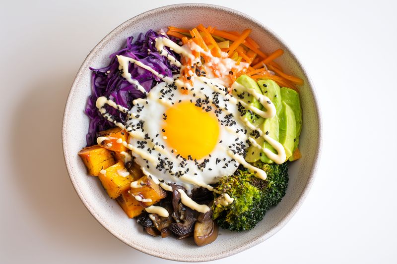 Bright healthy vegetable bowl with a sunny fried egg, avocado, roasted squash, broccoli, mushrooms, purple cabbage and shredded carrots, drizzled with creamy sauce and sesame seeds.
