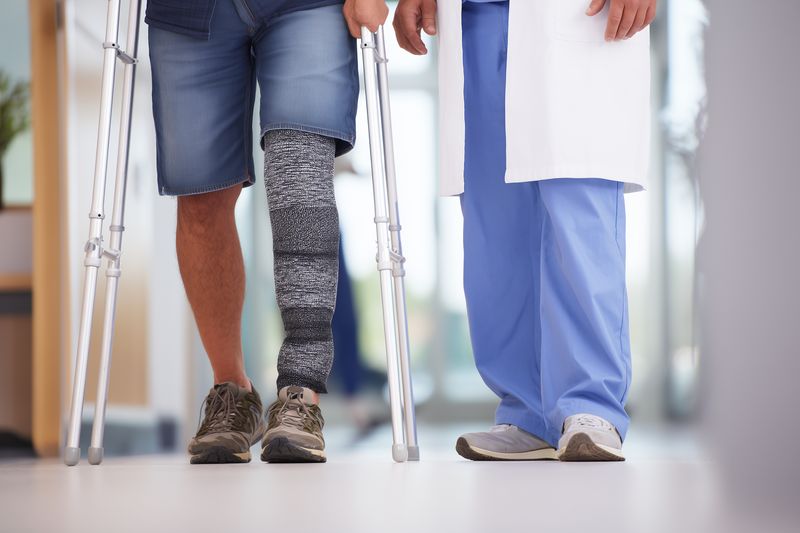 Male patient with a leg cast using crutches walks alongside a healthcare professional in a clinical corridor, receiving support during orthopedic recovery and mobility rehabilitation.