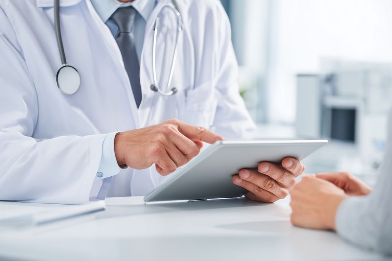 Doctor in white coat with stethoscope consulting a patient while using a digital tablet, close up of hands and medical workspace conveying professional healthcare interaction and technology.