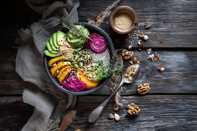 Colorful vegan Buddha bowl with sliced avocado, roasted squash, pickled beetroot, quinoa, chia and pumpkin seeds, microgreens, olive oil drizzle, styled on rustic wooden table.