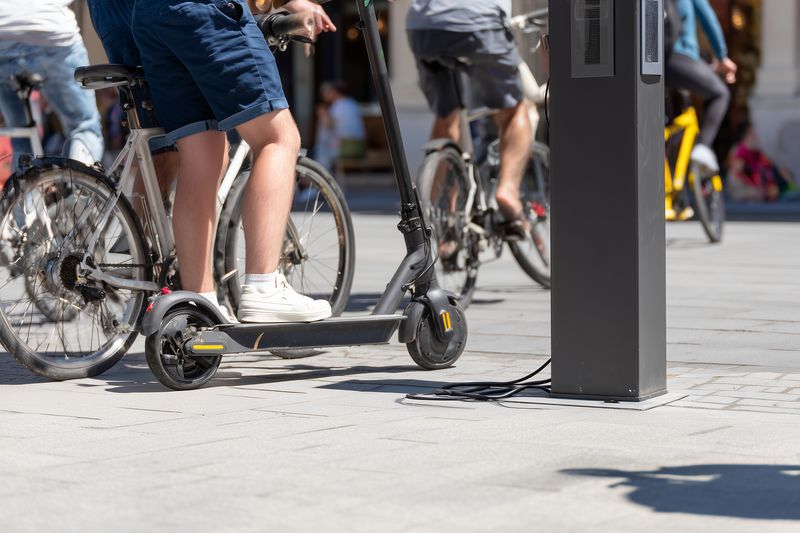 Electric scooter parked at a public charging station in a busy urban plaza alongside bicycles and pedestrians, capturing micro mobility and modern city transportation scene.