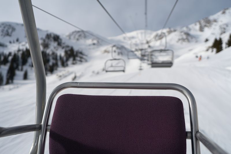 Empty chairlift seat with textured purple upholstery glides above snowy slopes toward distant mountain ridges, capturing quiet winter resort atmosphere and alpine landscape mood.