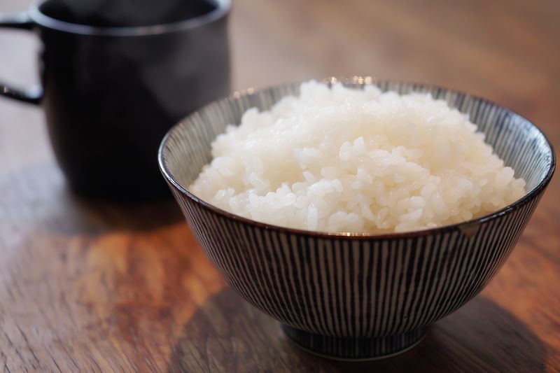 Close-up of steaming cooked white rice served in a ceramic bowl on a wooden table with a blurred cup in background, simple homey food presentation and warm natural light.