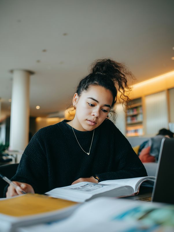 Young woman studying in a modern library or cafe, focused on reading a textbook and taking detailed notes with a laptop nearby, surrounded by warm lighting and a relaxed atmosphere.