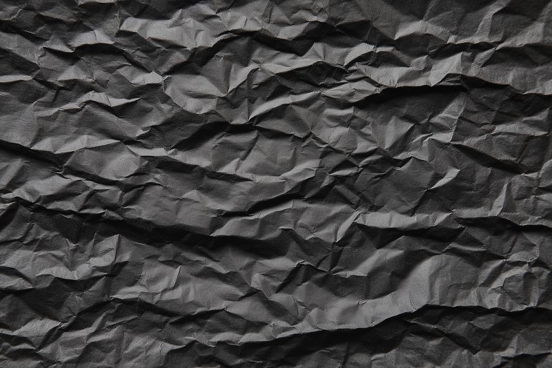 High contrast close-up of crumpled black paper texture showing intricate folds, creases and grain, creating dramatic light and shadow for backgrounds, design or artistic projects.