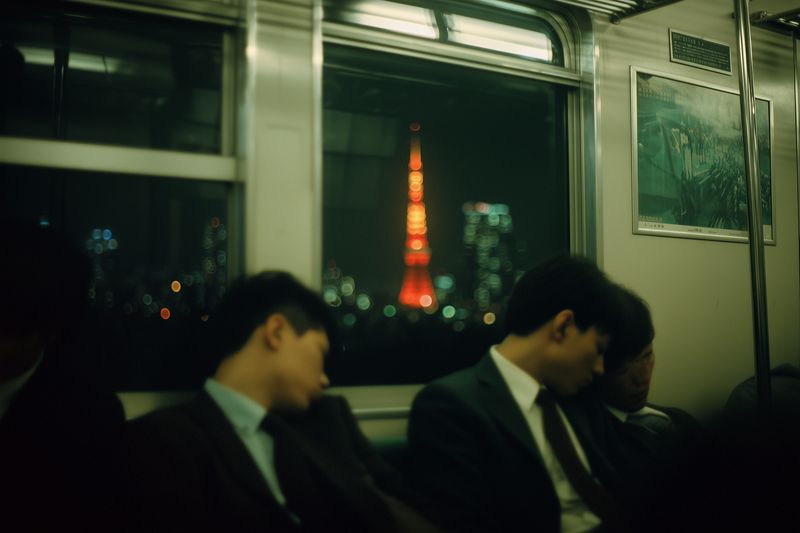 Grainy film photo of sleeping commuters on a night subway, blurred city tower glowing through the window, moody low-light atmosphere and candid urban solitude.