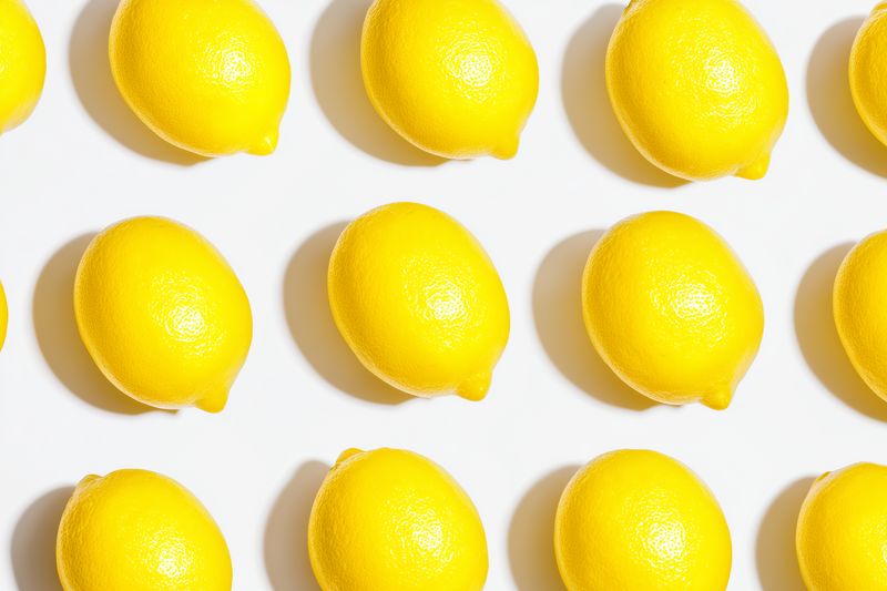 Bright yellow lemons arranged in a repeating grid on a clean white background, creating a vibrant minimalist pattern that emphasizes texture, color contrast and fresh citrus appeal.
