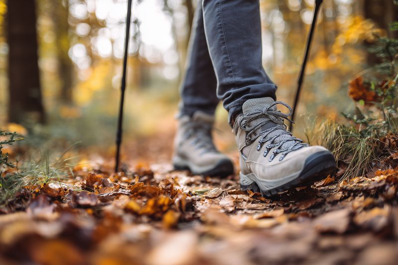 Close-up of hiking boots stepping through a leaf-covered forest trail in autumn, showing movement, rugged footwear and textured ground with warm seasonal colors.