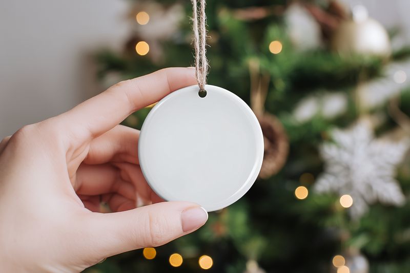A hand holds a blank round ceramic ornament against a blurred Christmas tree backdrop with warm bokeh lights, creating a cozy festive scene ready for personalization.