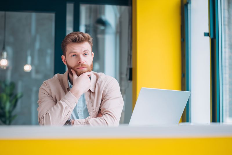 Young bearded man sitting at a laptop in a bright modern office, looking thoughtful and planning work, surrounded by contemporary decor and vibrant yellow accents for an energetic mood.