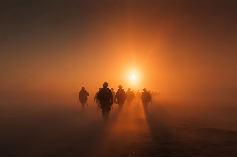 Silhouetted figures walk through dense orange fog toward a low glowing sun, casting long shadows across barren terrain and creating a dramatic, cinematic atmosphere of mystery.